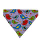 A triangular bandana-style pet collar or scarf made of lavender fabric, printed with a colorful pattern of cartoon birds in red, blue, green, and yellow on a light beige background.