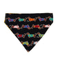 A triangular pet bandana or scarf laid flat on a light background, featuring a black base with a repeating pattern of dachshund dogs wearing colorful sweaters.